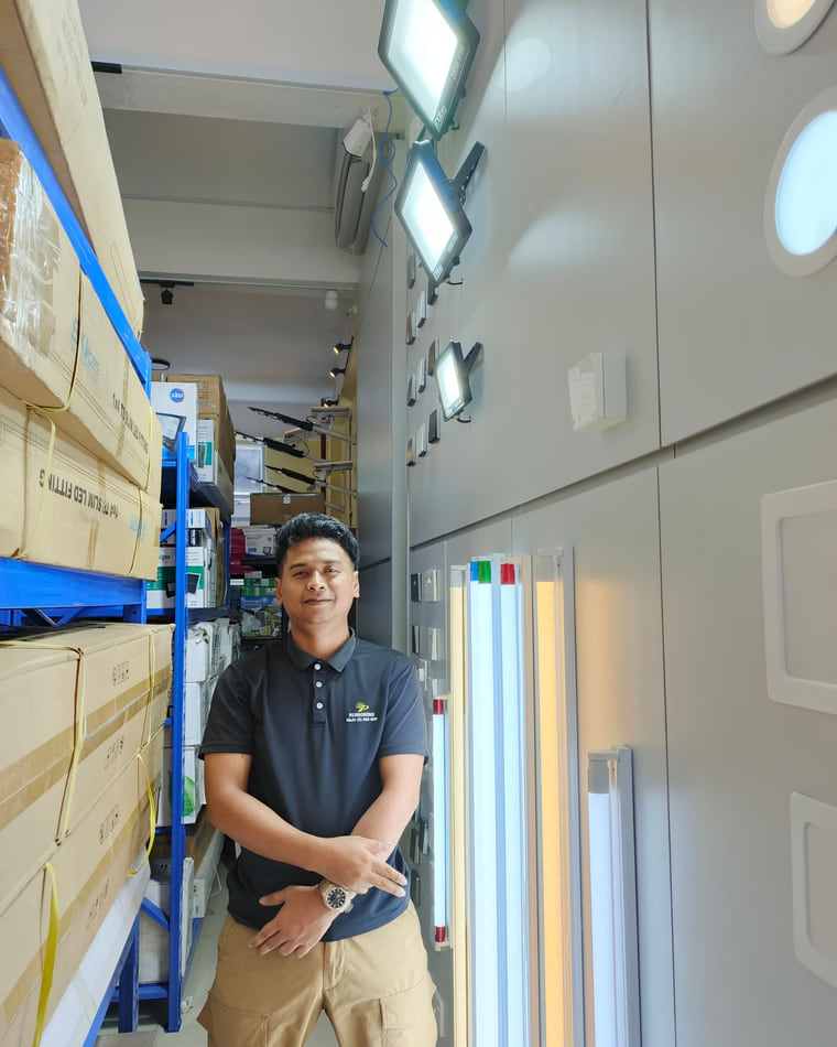 A showroom aisle at the Kolombong warehouse, racking on one side and a wall of lights on the other.